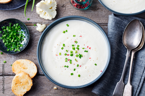 Cauliflower, potato cream soup with green onion in black bowl on grey wooden background