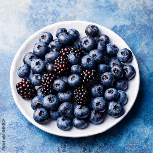 Blueberry and blackberry berries on a white plate on blue stone background. Top view.