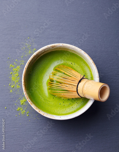 Matcha green tea cooking process in a bowl with bamboo whisk. Black slate background.