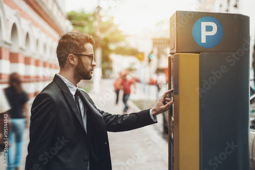 Young handsome businessman in glasses and the black business suit is paying his parking time using the automatic kiosk; the confident male employee is making payment with parking pay station terminal