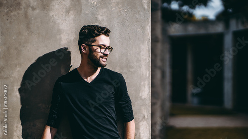 Portrait of smiling calm bearded hipster in glasses looking aside while standing in front of concrete wall outdoors on sunny day, strong shadow; with copy space zone for your advertising text or logo