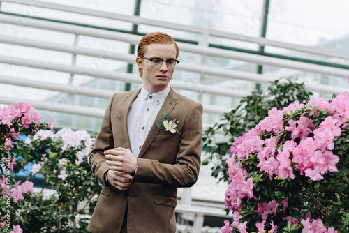 handsome stylish young redhead groom in eyeglasses looking away in botanical garden