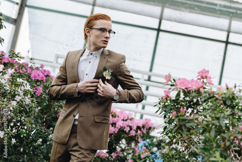 handsome stylish young redhead groom in eyeglasses wearing boutonniere and looking away in botanical garden