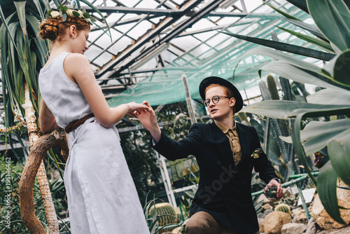 stylish young man in hat and eyeglasses making proposal to beautiful girl in botanical garden