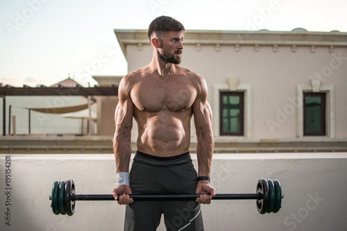 Shirtless muscular bearded man doing exercises with barbell outdoors.