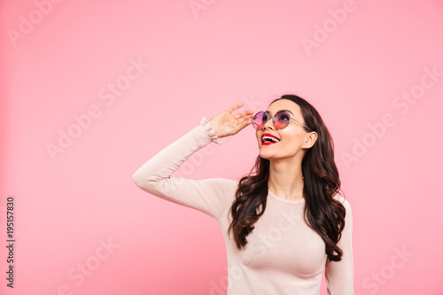 Portrait of elegant woman with red lips wearing round sunglasses smiling and looking upward on copy space, isolated over pink background
