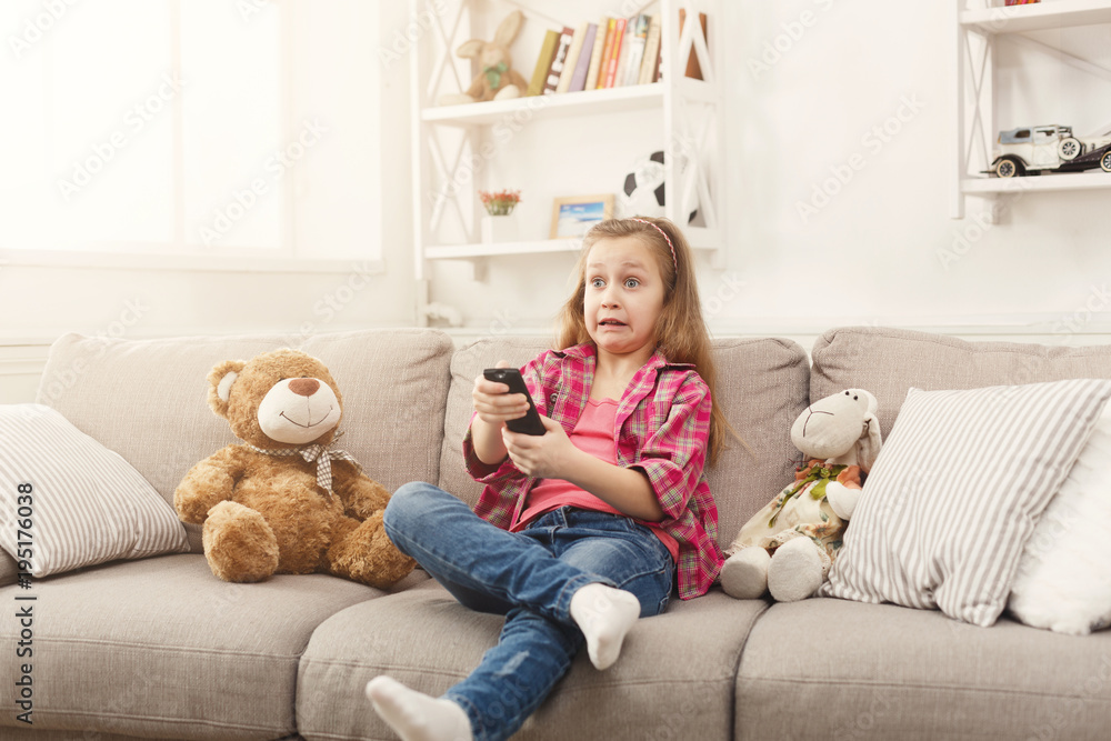 Confused casual little girl watching tv while sitting on sofa at home Confused casual little girl watching tv while sitting on sofa at home