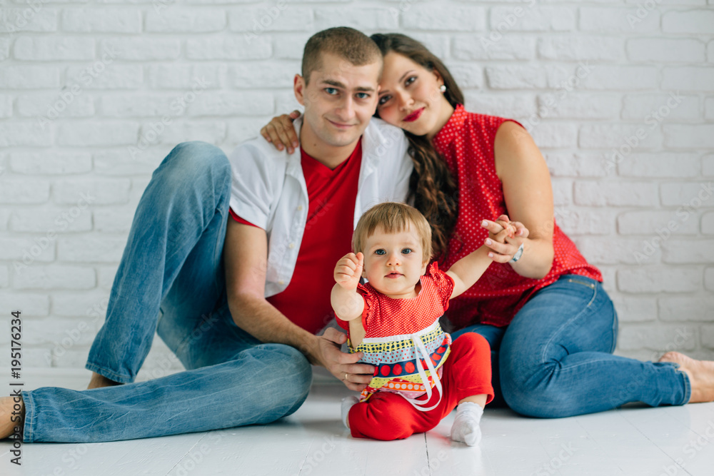 Fototapeta premium Young happy family sitting on the white floor and having fun. Parents with baby little girl in colorful dress. Man and woman in jeans. Brick wall. Mother father daughter.
