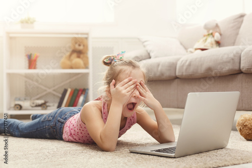 Scared casual little girl watching movie on laptop while lying on the floor at home © Prostock-studio