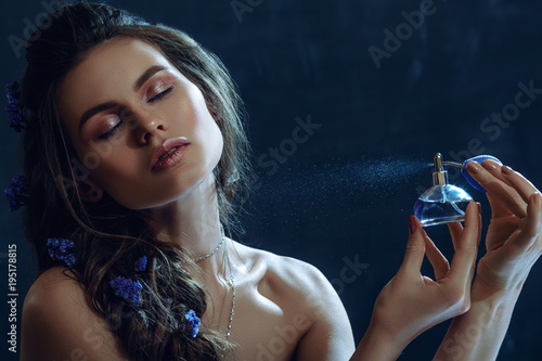 Close up studio portrait of young beautiful woman holding, spraying, using perfume in blue bottle. Model with nude makeup, greek braid hairstyle, posing on dark background. Copy, empty space for text photo