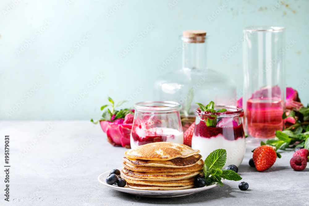 Stack of homemade pancakes served on plate with berries, mint, glass jars of yogurt, bottle of lemonade, fruit salad in pink dragon fruit over grey texture table. Copy space Stack of homemade pancakes served on plate with berries, mint, glass jars of yogurt, bottle of lemonade, fruit salad in pink dragon fruit over grey texture table. Copy space