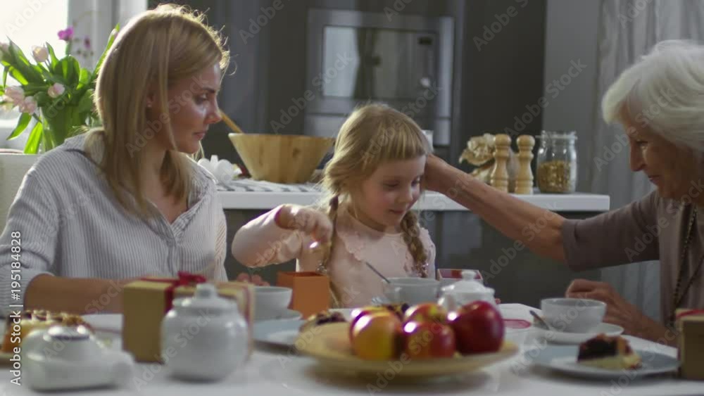 Cute little girl sharing cookies with mother and grandmother and kissing them while having family dinner Cute little girl sharing cookies with mother and grandmother and kissing them while having family dinner
