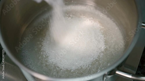 Close up of sugar being strewed into steel bowl with water on the bottom for cake cooking in the kitchen. Candy mass is melting slowly and mixing with liquid and some white sand is above the beverage