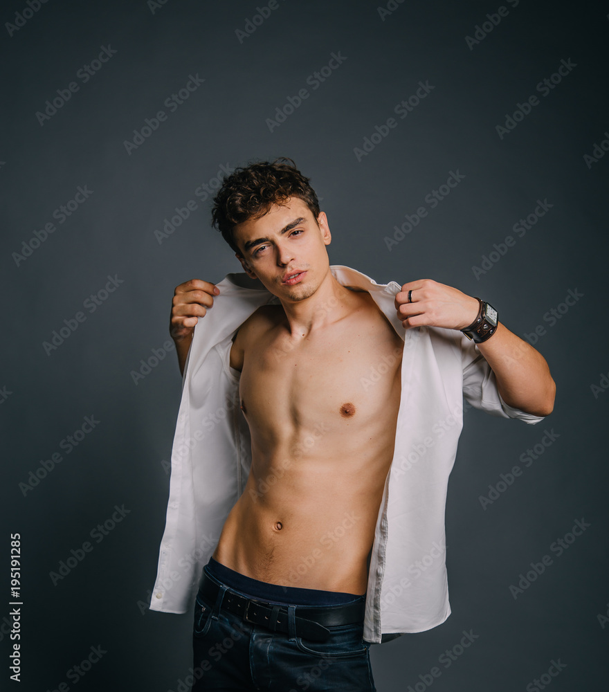 A young, handsome guy posing with an unbuttoned, white shirt, showing a sporty body, muscles, relief, pumped-up body. Background gray. Studio shooting, minimal, natural retouching