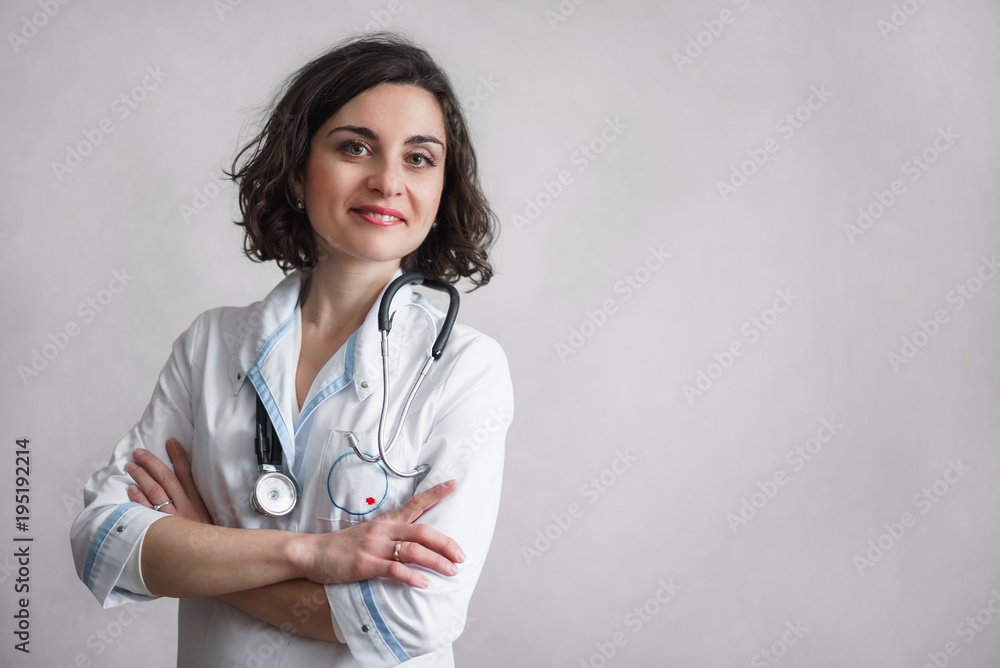 Cute smiling woman with dark curly hair in white medical robe with blue finish stands against a gray wall and looks at the camera. On the neck of a stethoscope. Cute smiling woman with dark curly hair in white medical robe with blue finish stands against a gray wall and looks at the camera. On the neck of a stethoscope.