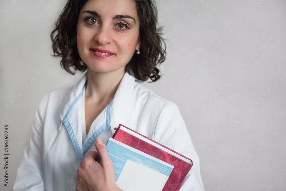 Cute smiling woman with an attentive serious look in white medical gown stands against a gray wall and looks at the camera. Holding a books. Cute smiling woman with an attentive serious look in white medical gown stands against a gray wall and looks at the camera. Holding a books.