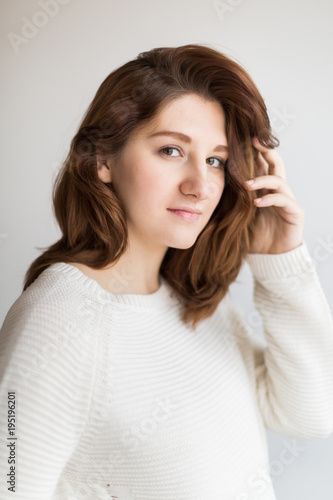 style, make up, beauty concept. close up of pretty face of young woman, she is holding her hairs with her small delicate hand, she is wearing clean white sweater