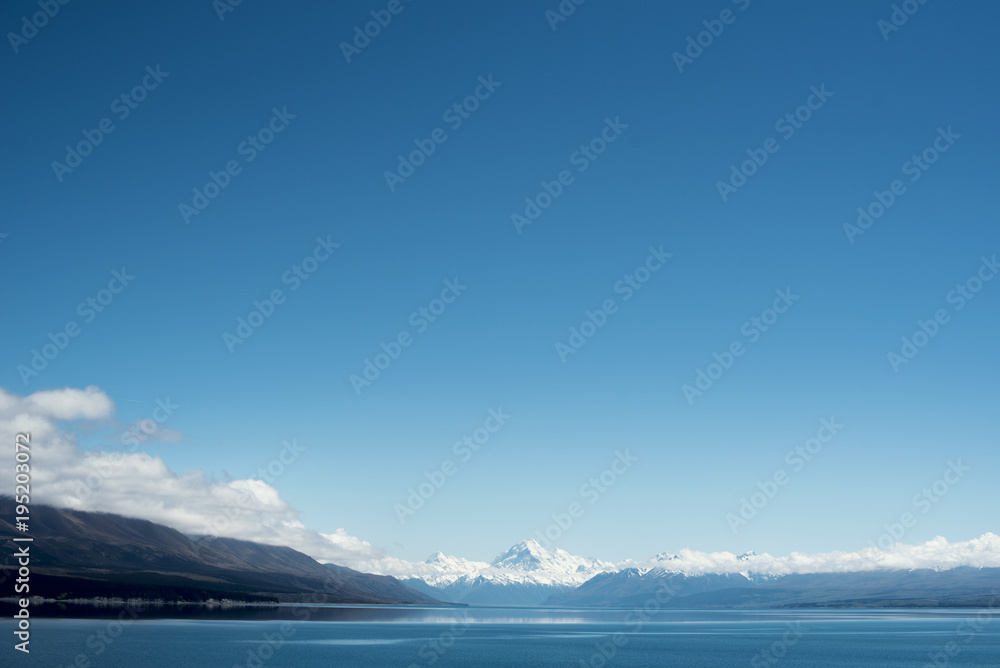 Paisaje de picos de montañas nevados con cielo azul y nubes frente un lago y valle con árbol en el Monte Cook, Nueva Zelanda.