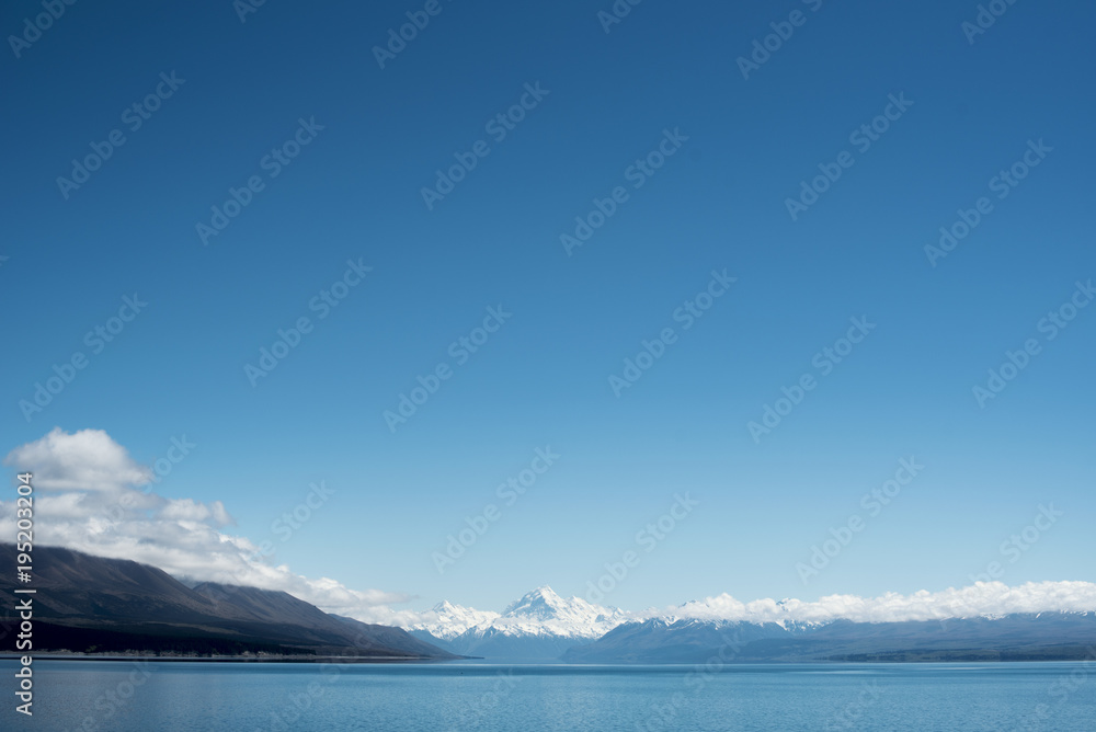 Paisaje de picos de montañas nevados con cielo azul y nubes frente un lago y valle con árbol en el Monte Cook, Nueva Zelanda.
