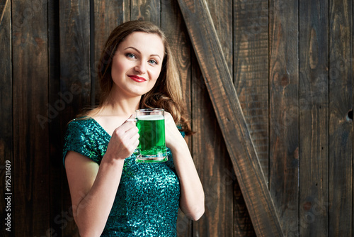 Smiling woman holding mug of green beer for St. Patricks Day celebrations on wooden background, copy space. Girl in emerald sequins dress holding glass of beer in pub. Greeting card, March 17