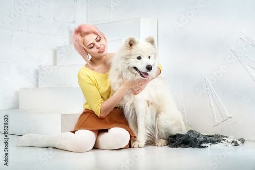 attractive young girl with short blond hair in a yellow sweater and skirt hugs her beloved pet - a dog breed Samoyed