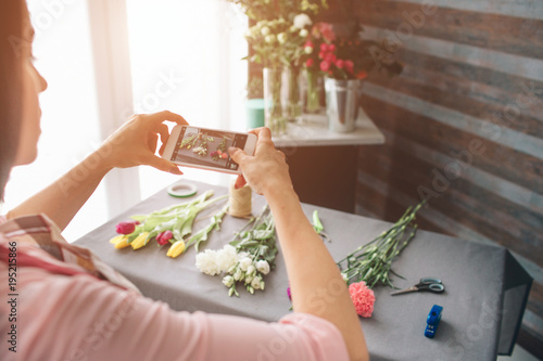 Female florist at work: pretty young dark-haired woman making fashion modern bouquet of different flowers. Women working with flowers in workshop. She is photographing flowers on a smartphone.