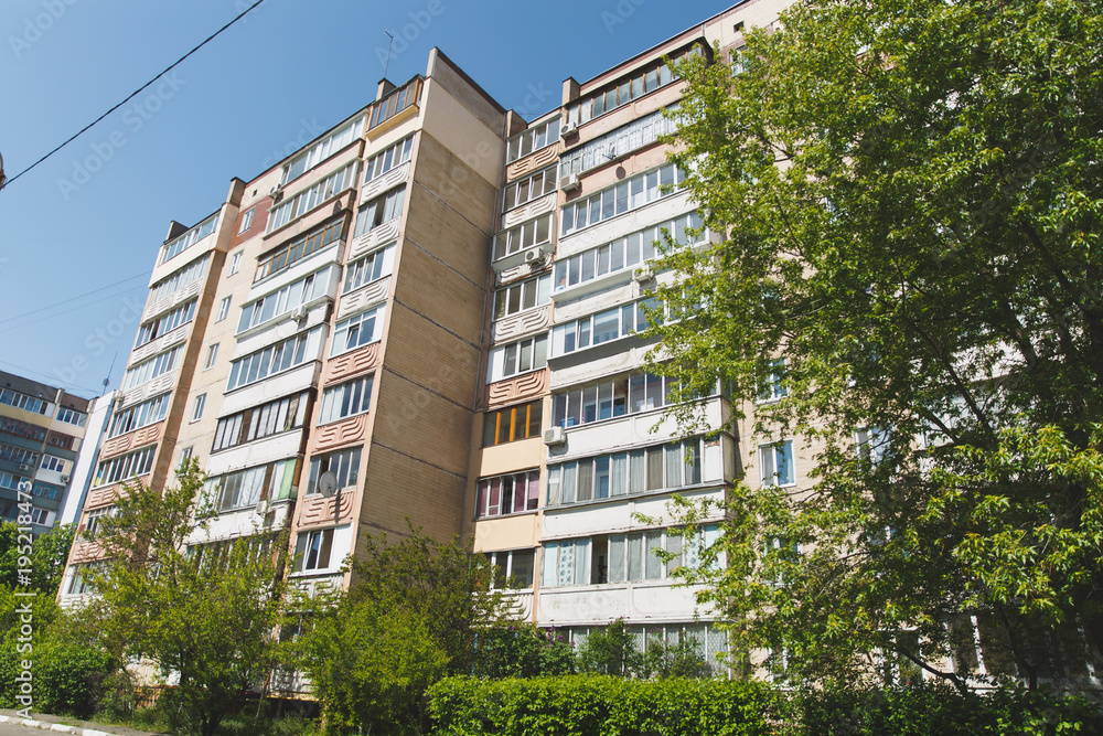 The facade of multi-storey high-rise Soviet-built house surrounded by green trees and bushes on a background of clear blue sky. Architecture, history