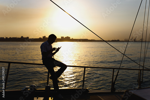 Silhouettes a young man in a suit who rests on a yacht, leans on a railing and typing on the screen of his smartphone on a sea walk against a beautiful sunset