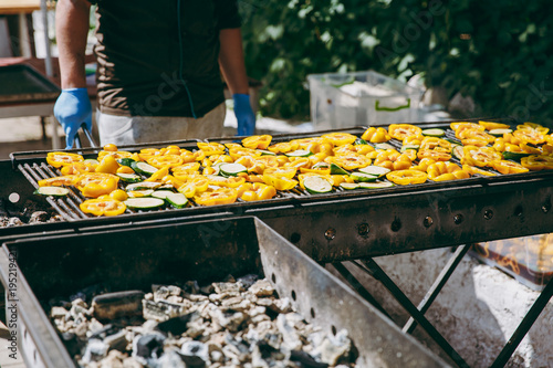 A man in gloves with tongs cooks and fries spread on a large grill bright tasty vegetables yellow sweet Bulgarian pepper and green zucchini outdoors in the garden at the barbecue party for guests