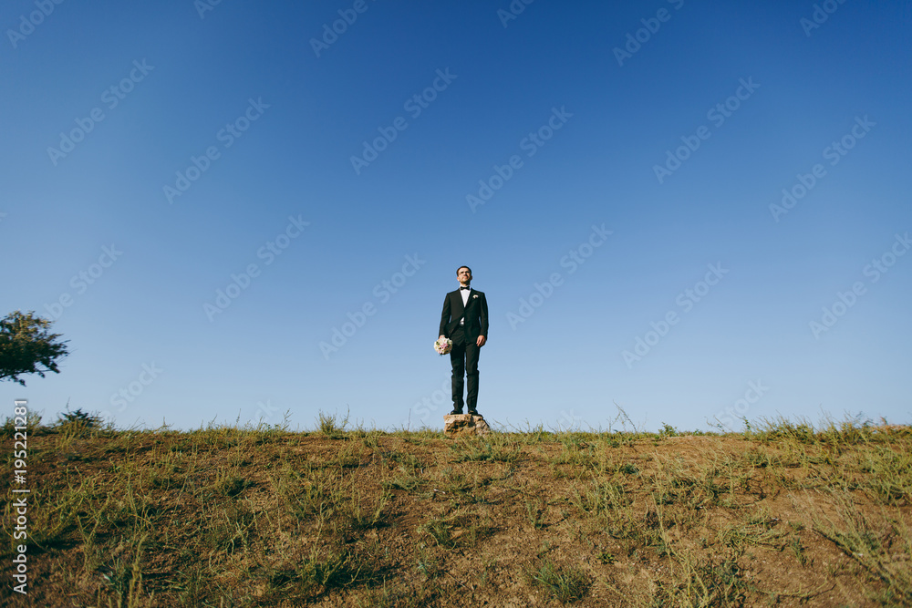 Beautiful wedding photosession. Handsome bridegroom in a black suit and white shirt with a bouquet of the bride stands on a stone on walk around the big green field against blue sky background