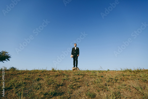Beautiful wedding photosession. Handsome bridegroom in a black suit and white shirt with a bouquet of the bride stands on a stone on walk around the big green field against blue sky background