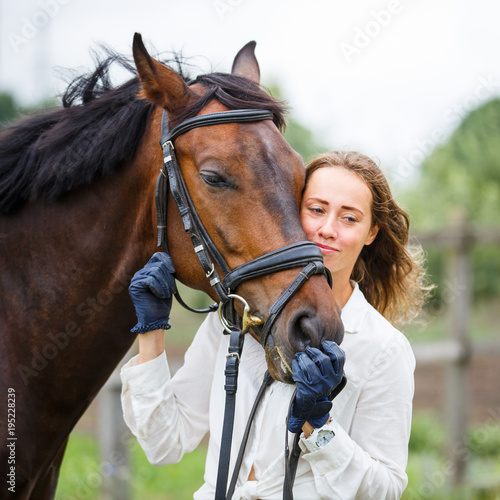Young happy rider woman with her bay horse. Portrait of smiling girl with her stallion in paddock on summer day © skumer