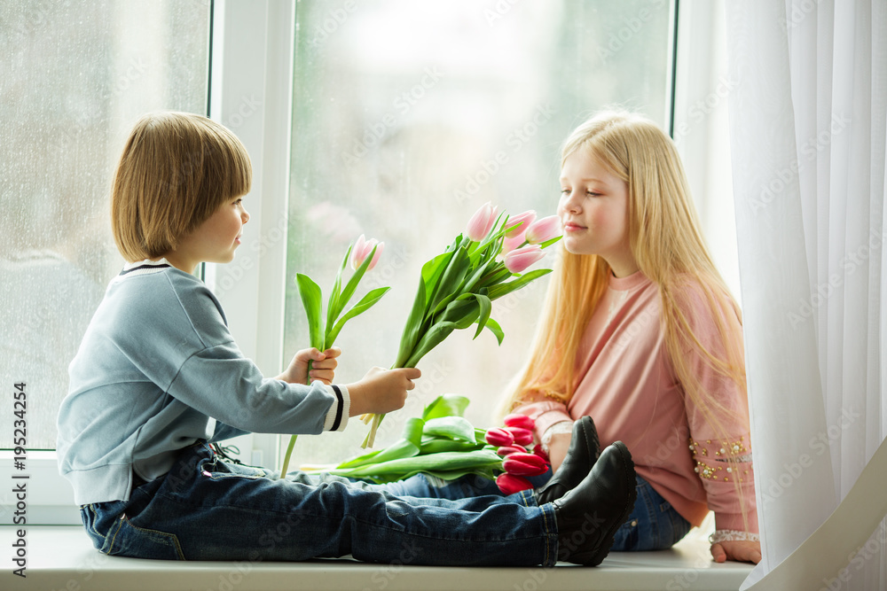 Pretty lovely kids are sitting on window sill , happy little boy giving a cute girl bouquet of pink tulip flowers. Easter concept Pretty lovely kids are sitting on window sill , happy little boy giving a cute girl bouquet of pink tulip flowers. Easter concept