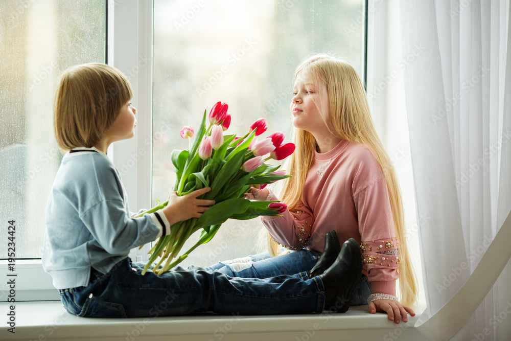 Pretty lovely kids are sitting on window sill , happy little boy giving a cute girl bouquet of pink tulip flowers. Easter concept Pretty lovely kids are sitting on window sill , happy little boy giving a cute girl bouquet of pink tulip flowers. Easter concept