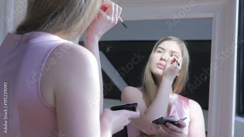 Beautiful blonde woman doing makeup in front of mirror. Smiling woman in pink dress behind mirror. Attractive girl doing makeup. Fashion and cosmetics concept, lips and eyes makeup.  