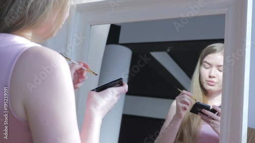 Beautiful blonde woman doing makeup in front of mirror. Smiling woman in pink dress behind mirror. Attractive girl doing makeup. Fashion and cosmetics concept, lips and eyes makeup.  
