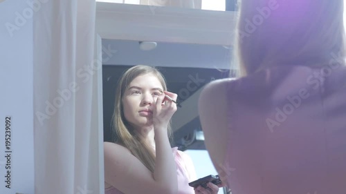 Beautiful blonde woman doing makeup in front of mirror. Smiling woman in pink dress behind mirror. Attractive girl doing makeup. Fashion and cosmetics concept, lips and eyes makeup.  