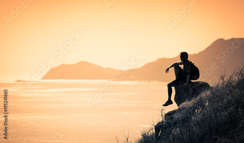 Young thoughtful man sitting on edge of mountain watching the sunrise. 