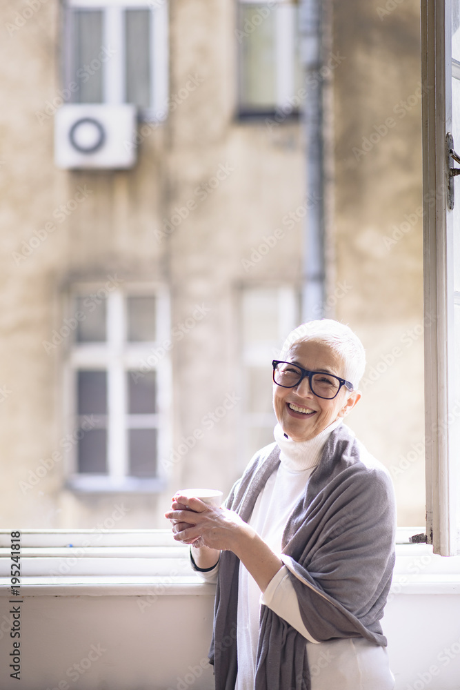 Senior elder woman enjoying her cup of coffee in the morning, standing next to a big home window Senior elder woman enjoying her cup of coffee in the morning, standing next to a big home window