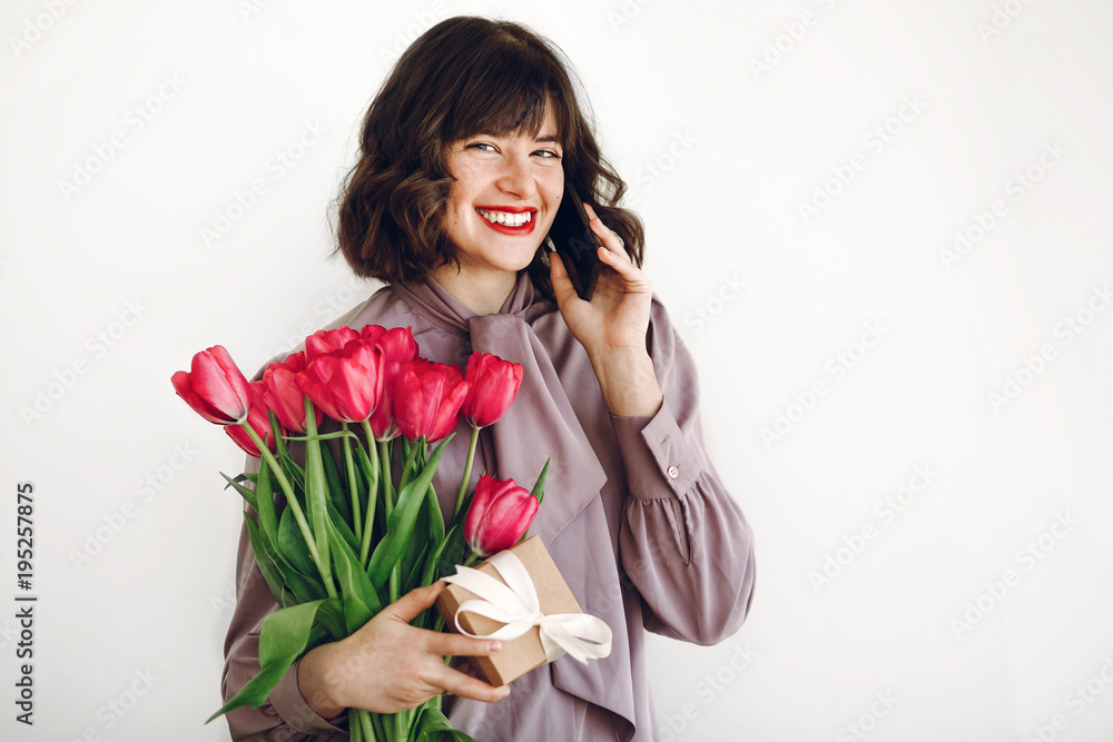 happy stylish girl talking on phone and holding pink tulips and gift box with ribbon on white background. happy mothers or womens day concept. young hipster woman smiling with flowers happy stylish girl talking on phone and holding pink tulips and gift box with ribbon on white background. happy mothers or womens day concept. young hipster woman smiling with flowers