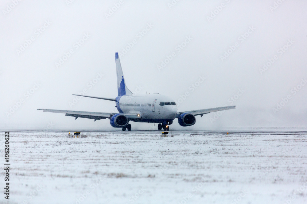 Obraz premium Airliner on runway in blizzard. Aircraft during taxiing at heavy snow. Passenger plane in snow at airport. Modern twin-engine passenger airplane taxiing for take off at airport during snow blizzard