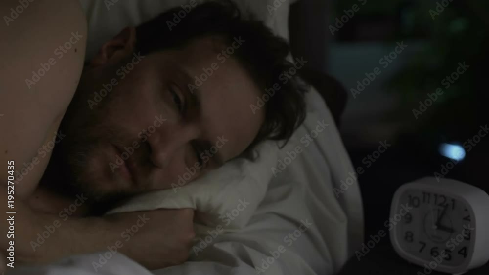 Man in his 40s suffering insomnia, turning in bed and looking angrily at clock Man in his 40s suffering insomnia, turning in bed and looking angrily at clock