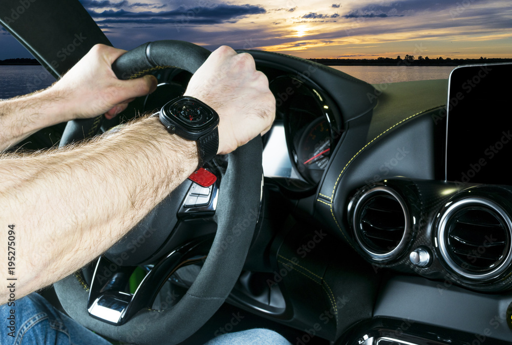 Male hands holding car steering wheel. Hands on steering wheel of a car driving near the lake. Man driving a car inside cabin. Multimedia system and dashboard. Speedometer, tachometer