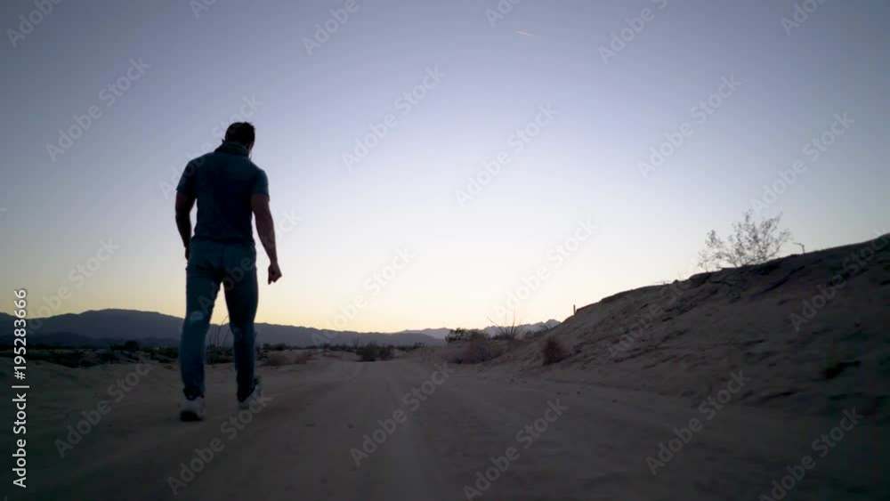 Camera at ground level and off to the side, a young well built man walks along a sandy road through the desert at sunrise. Camera at ground level and off to the side, a young well built man walks along a sandy road through the desert at sunrise.