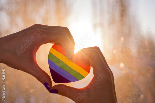 lgbt rights concept, hands in the form of heart holds a heart painted like a LGBT flag, silhouetted against sun photo