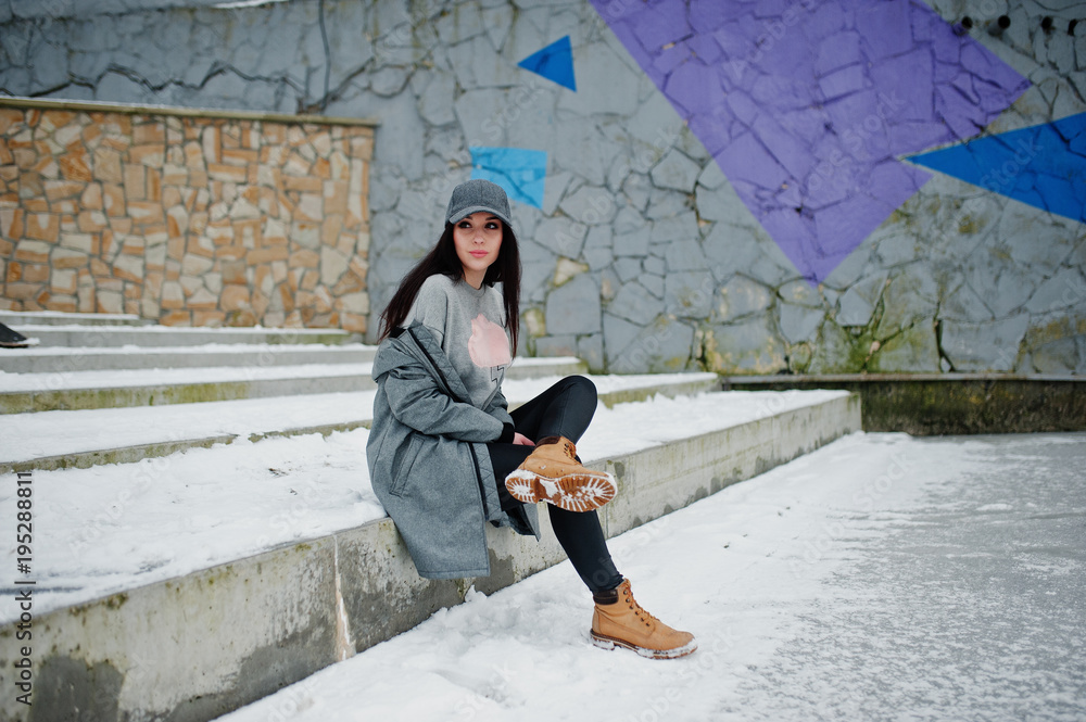 Stylish brunette girl in gray cap, casual street style on winter day against colored wall. Stylish brunette girl in gray cap, casual street style on winter day against colored wall.