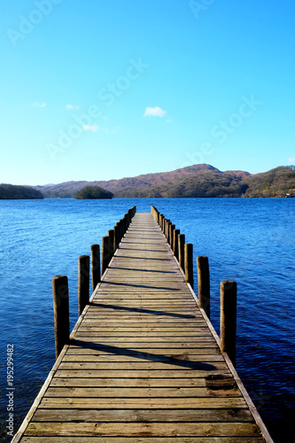 very long beautiful wooden jetty, jutting out from the centre of the image into a calm blue lake with hills of forest and meadows in background