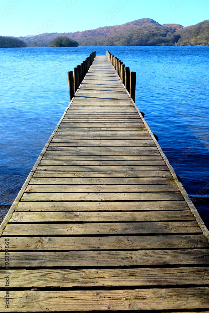 Obraz premium long wooden symetrical beautiful wooden jetty, jutting out from the centre of the image into a calm blue lake with hills of forest and meadows in background