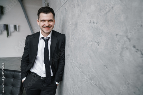 Quadro em tela Portrait of a young attractive man in a black suit and tie with a smile and confident look against a gray wall background in a trendy loft style