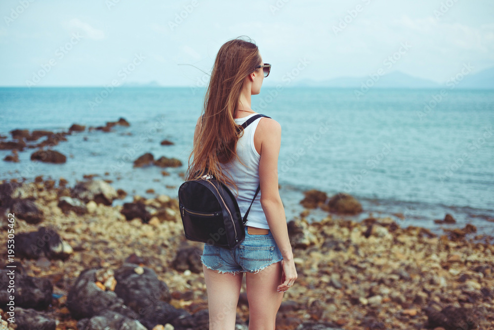 back view of young woman in sunglasses with backpack looking at ocean